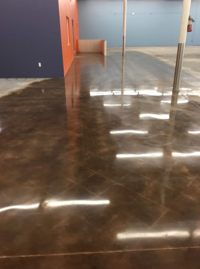 A large room with a high-shine marbled brown concrete floor, showing reflections of ceiling lights and colored walls, with a fire extinguisher mounted on a column, giving the space a functional yet polished appearance