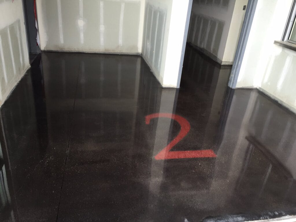 A corner of a room with glossy dark flooring that reflects the surrounding walls and a large red number '2' painted on the floor. The walls are light-colored with some patchy areas indicating possible ongoing work or repairs. Multiple doorways and a window frame suggest this is an interior space within a larger building. The presence of the number on the floor could imply a wayfinding element in a commercial or industrial environment.