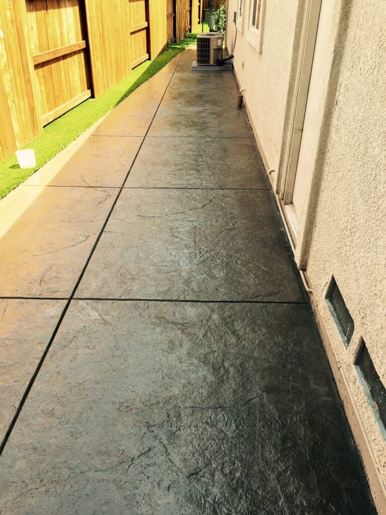 A narrow walkway with large stone tiles runs alongside a building with stucco walls, leading towards a wooden fence and an HVAC unit, with a strip of artificial grass on the opposite side.