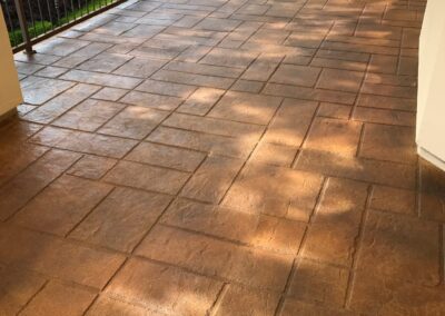 Covered patio with terracotta-colored stamped concrete flooring, leading to an ornate wrought iron railing overlooking a lush green landscape, invoking a sense of warmth and serene outdoor living.