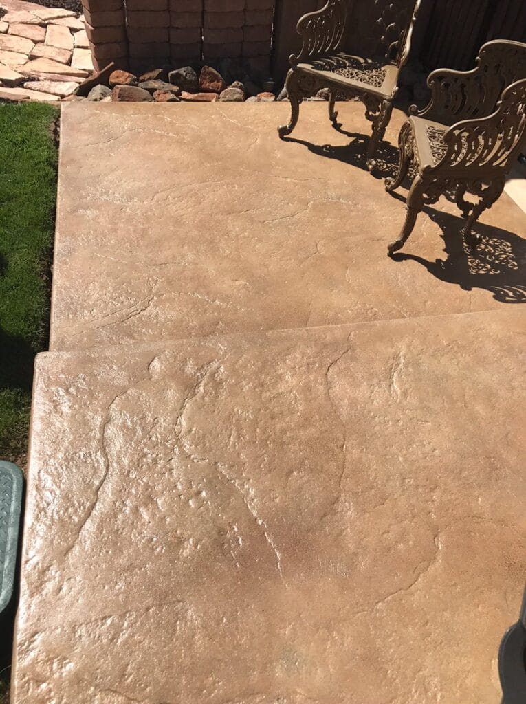 An outdoor patio with textured concrete paving extends towards an ornate metal bench, with a neatly manicured lawn and a brick retaining wall with rocks at the edge, suggesting a well-maintained garden space.