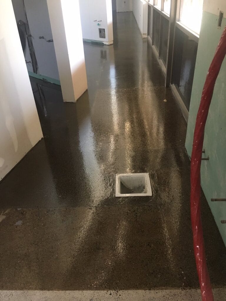 A narrow walkway with large stone tiles runs alongside a building with stucco walls, leading towards a wooden fence and an HVAC unit, with a strip of artificial grass on the opposite side.