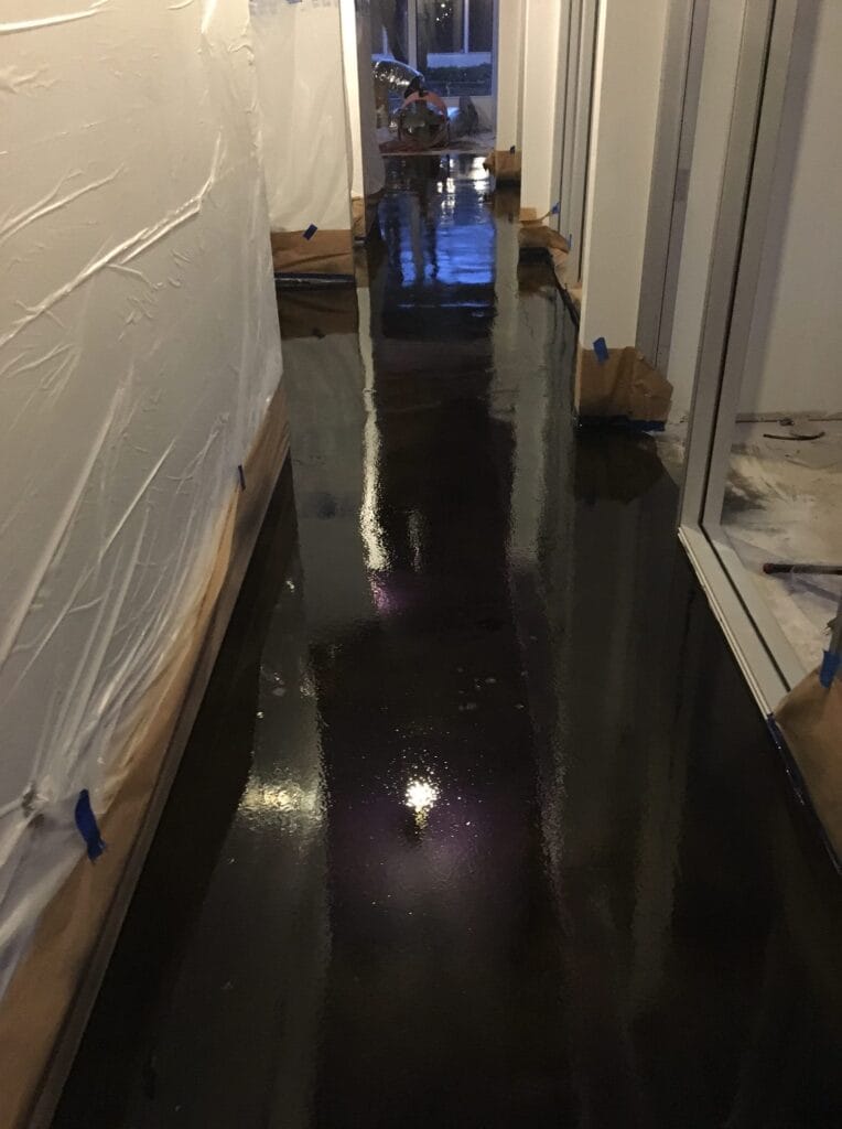 A narrow walkway with large stone tiles runs alongside a building with stucco walls, leading towards a wooden fence and an HVAC unit, with a strip of artificial grass on the opposite side.