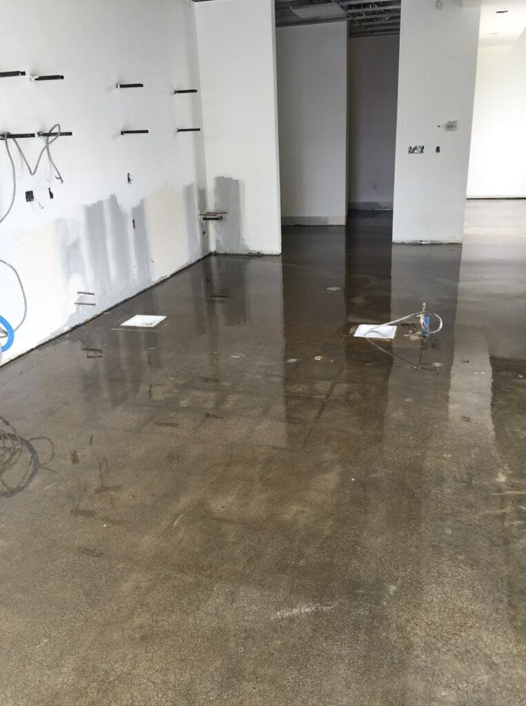 An unfinished room with concrete floors showing water puddles and reflections, with partially painted walls and exposed cabling, suggesting recent construction or repair work.