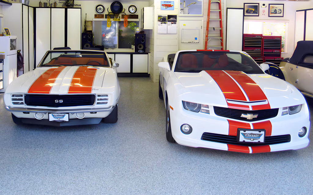 Two Chevrolet Camaros inside a brightly lit garage, facing forward. On the left is a classic 1969 Camaro SS with white paint and orange racing stripes, while on the right is a modern white Camaro with red stripes. The garage is well-organized with shelves, speakers, and automotive decor.