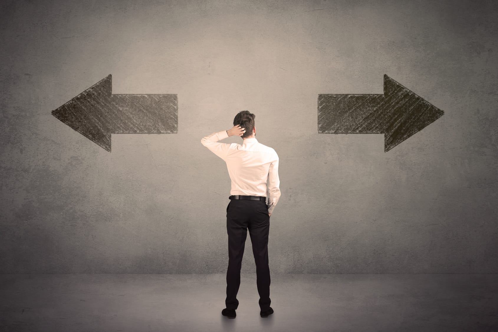 A man in business attire stands in front of a concrete wall, looking upwards and scratching his head in a gesture of contemplation or decision-making. Behind him, two large, chalk-drawn arrows point in opposite directions, symbolizing a crossroad or a need to make a choice between two different paths or options.