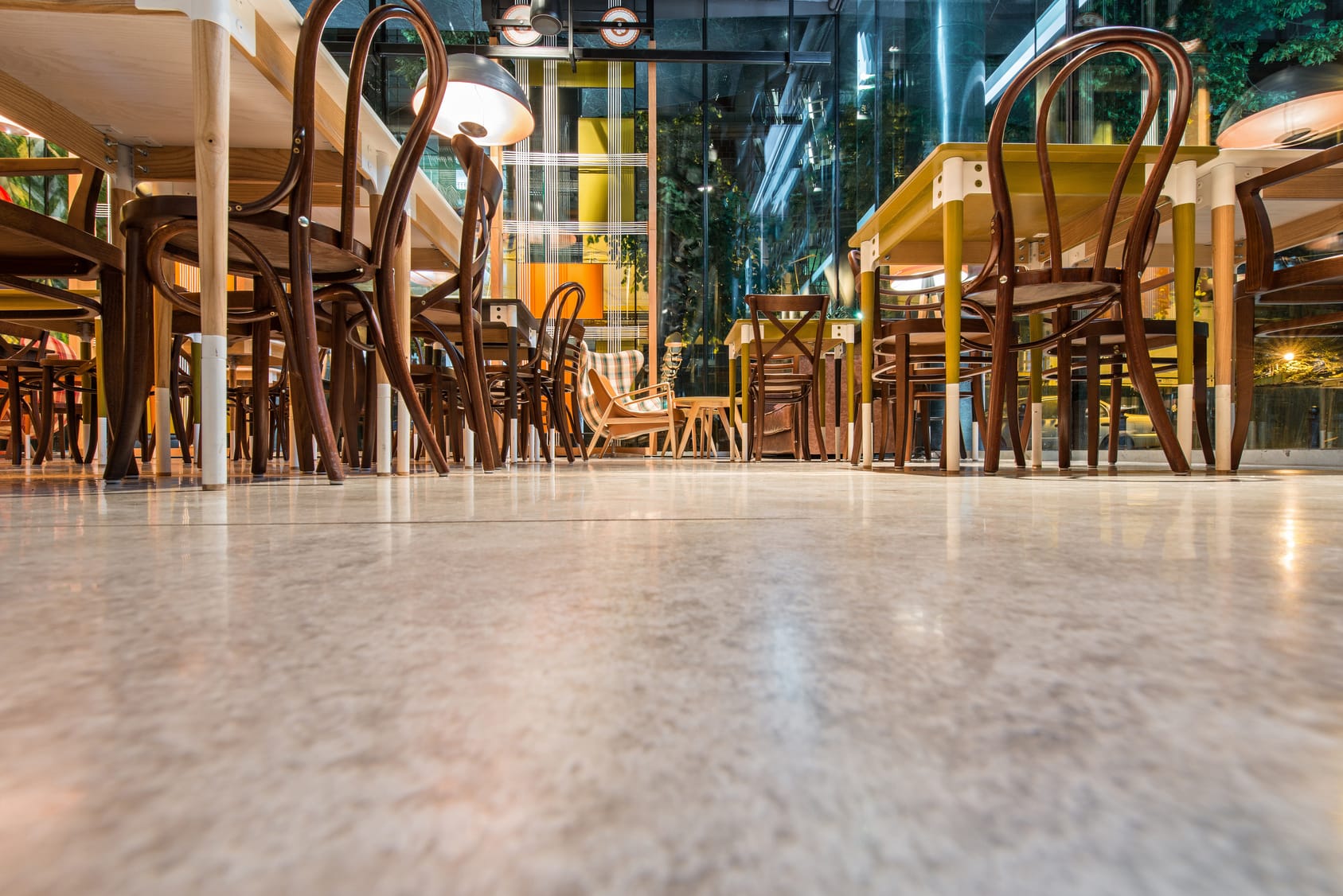 A ground-level view inside a modern cafe with an array of wooden chairs and tables. The furniture is neatly stacked, indicating the cafe is either closed or not yet open for customers. The polished stone floor reflects the ambient lighting, contributing to the establishment's clean and inviting atmosphere. The glass walls provide a glimpse of the urban setting outside.