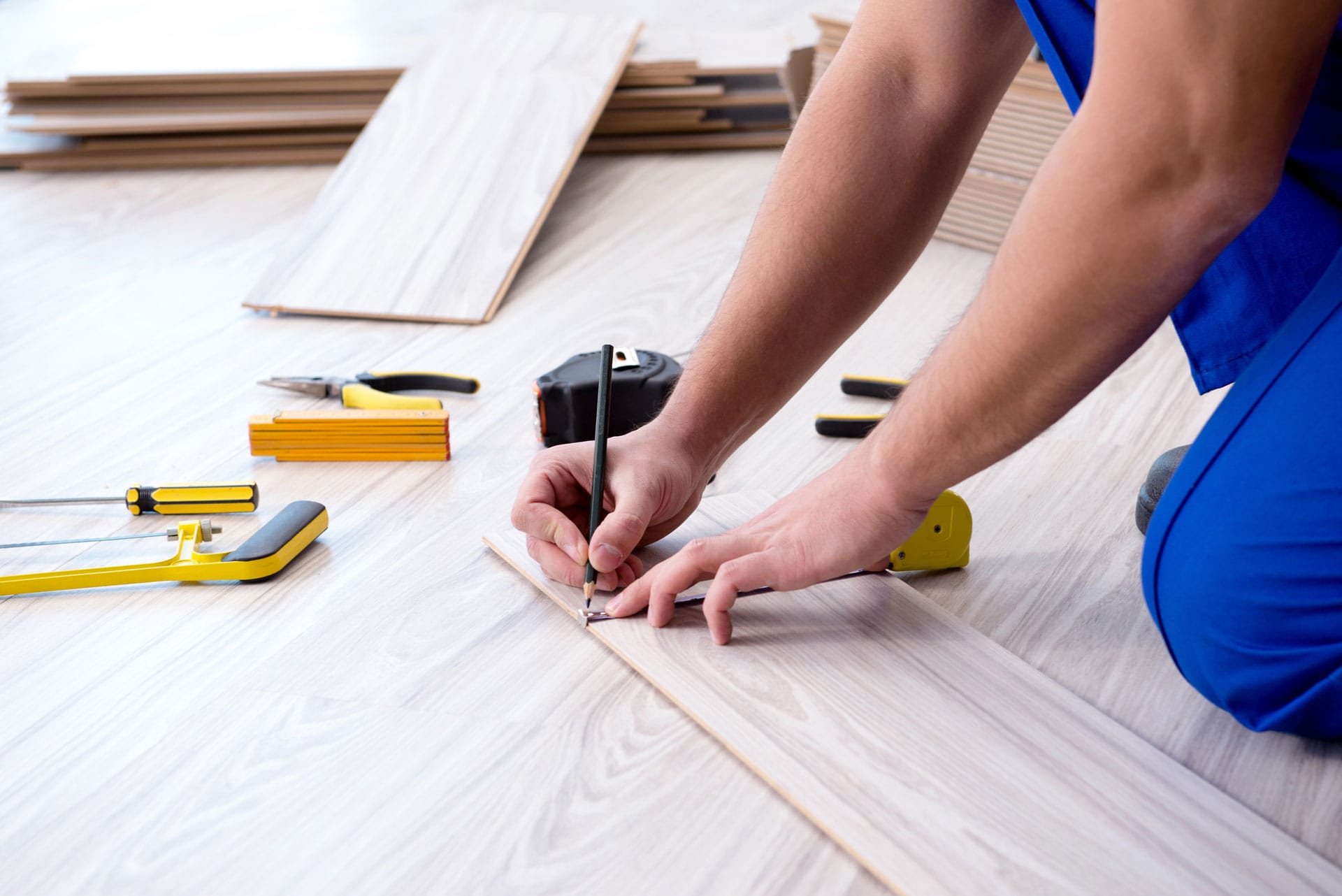 A person in blue attire is installing laminate flooring, measuring and marking the wooden plank to fit. Flooring installation tools, such as a handsaw, clamps, a tape measure, and other equipment, are scattered around the work area. The focus is on the hands performing a precise measurement, indicating skilled manual work.