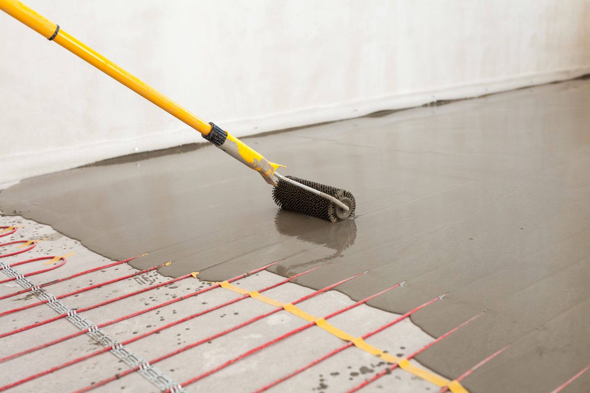 Application of a self-leveling compound on a concrete floor using a spiked roller attached to a yellow pole. The floor has underfloor heating pipes laid out in a grid pattern, visible through the semi-liquid layer of the compound. This image represents a stage in preparing a floor for final surface installation.