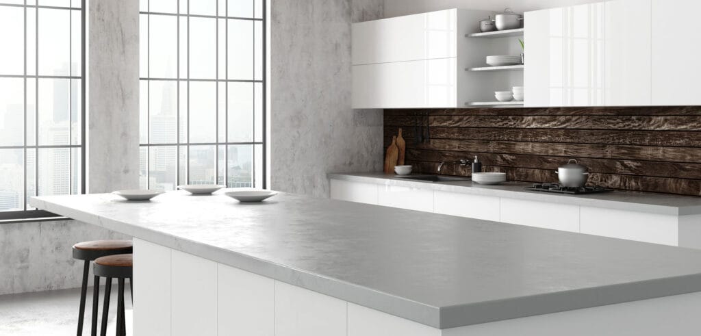 A modern kitchen interior with a large window providing a cityscape view. The kitchen features a minimalist design with a white concrete countertop, bar stools, and a wooden backsplash. Open shelving holds neatly arranged dishes and kitchenware. Natural light fills the room, creating a bright and airy atmosphere.