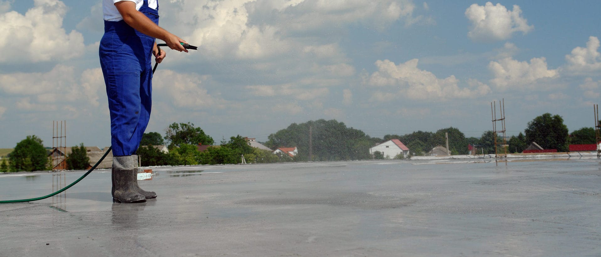A worker in a blue uniform and rubber boots using a hose to clean a large concrete surface on a rooftop, with a village and cloudy sky in the background.