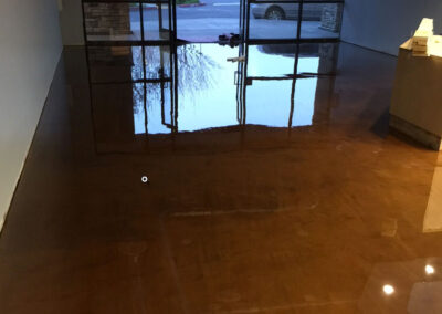 Interior space with a glossy, caramel-colored epoxy floor reflecting the glass entrance doors and the outdoor view, creating a mirror-like effect.