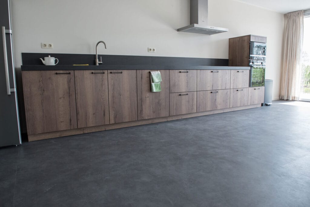 Modern kitchen interior with long wooden cabinets and black countertop, featuring a stainless steel sink and hanging range hood, with a minimalist grey epoxy floor."