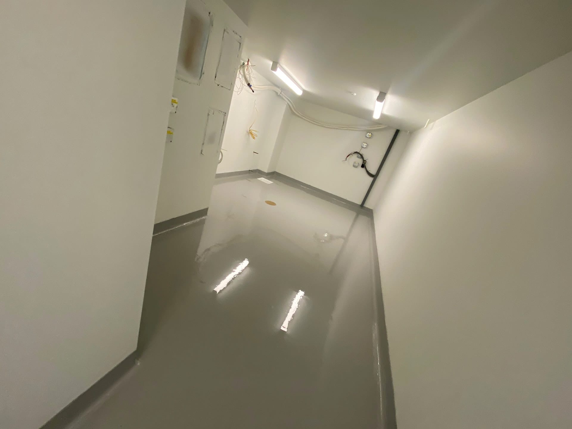 Glossy Light Gray Epoxy Coating An interior view of a room under construction with freshly applied grey epoxy flooring that reflects the ceiling lights, surrounded by white walls with exposed electrical wiring and fixtures.
