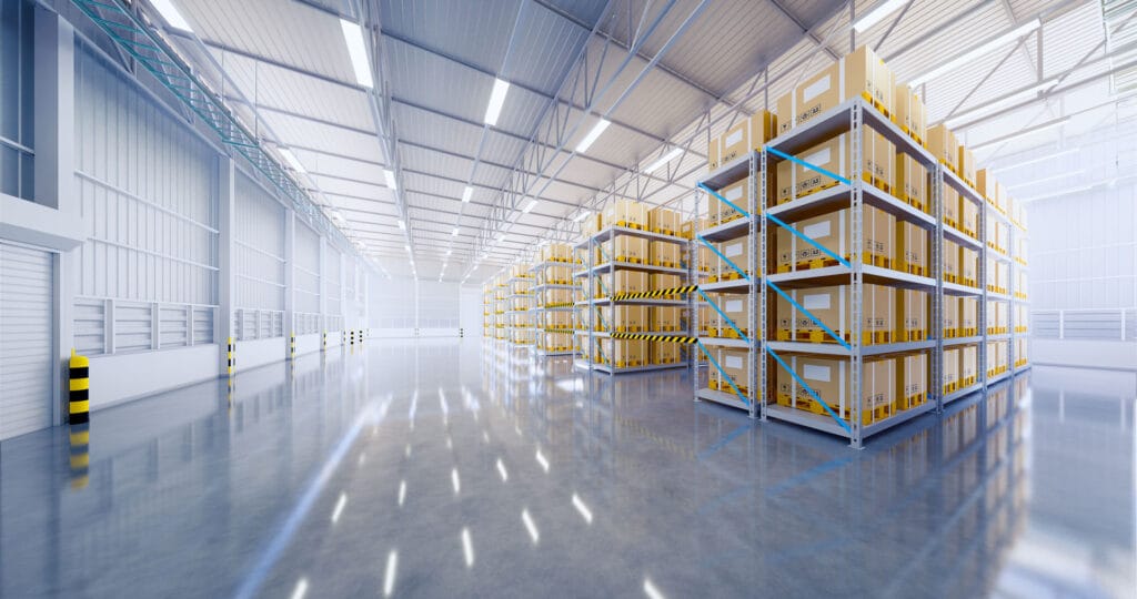 Spacious and modern warehouse interior with high ceilings and rows of tall metal shelving units storing numerous yellow and brown boxed goods. The floor is glossy, reflecting the bright LED lights above, and the warehouse has a clean, organized appearance.