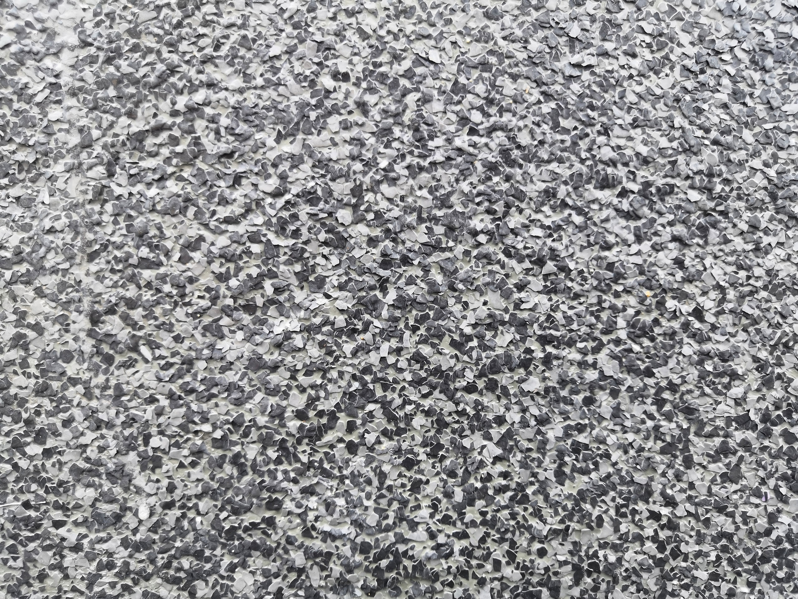 Close-up of a rough-textured surface with a speckled pattern of small, irregular black, gray, and white flakes, resembling a pebble dash or terrazzo floor finish.
