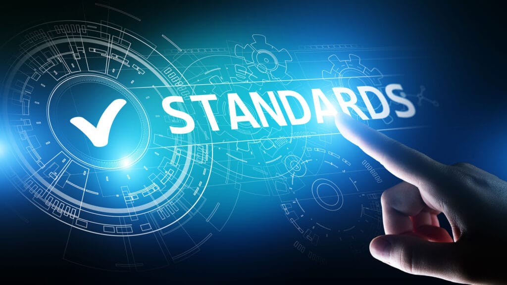Close-up of a hand pointing toward the word "STANDARDS" on a digital interface with futuristic blue HUD (heads-up display) elements, including gears and circuit-like graphics. A large white checkmark is shown inside a glowing circle, symbolizing approval or compliance.