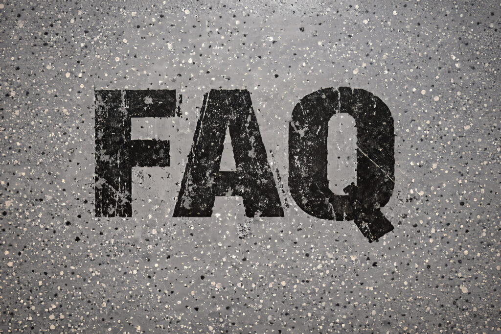 Overhead view of a gray epoxy garage floor with black, distressed ‘FAQ’ lettering embedded directly into the coating, showing a gritty, industrial texture.