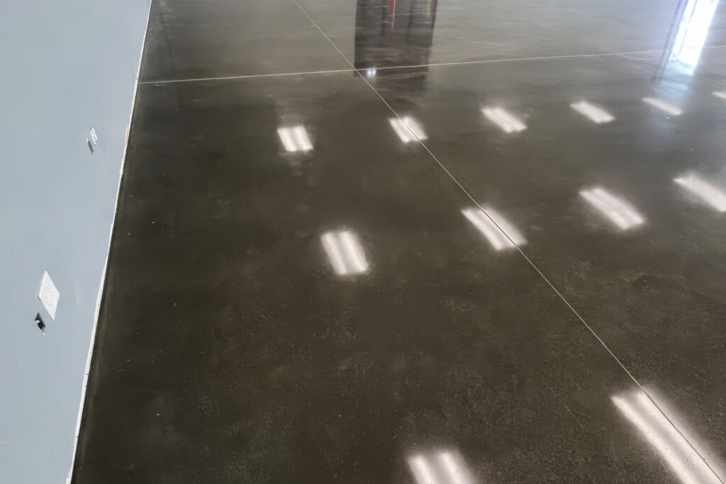 Smooth polished concrete floor near a gray wall, reflecting ceiling lights across its glossy surface.