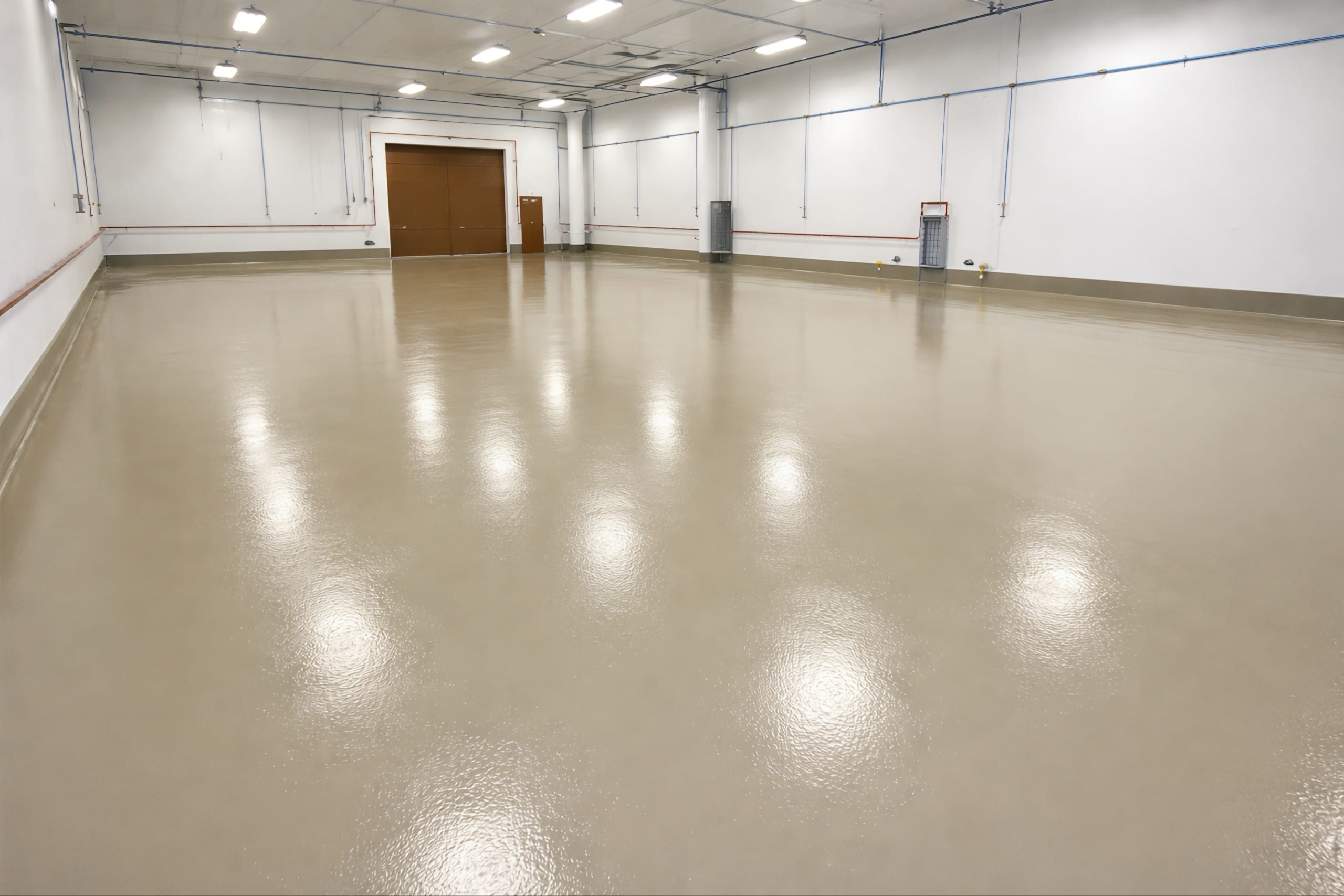 Spacious industrial room with a smooth, glossy urethane-coated floor in a neutral beige tone, reflecting overhead lights, with white walls, exposed piping, and a large brown door in the background.